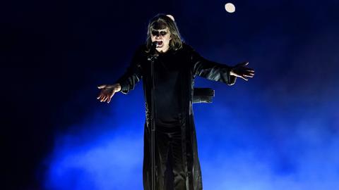 Der Heavy-Metal-Sänger Ozzy Osbourne steht auf einer Konzertbühne. Er trägt einen bodenlangen schwarzen Mantel und singt mit ausgebreiteten Armen.