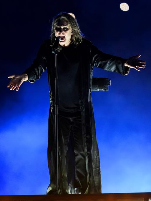 Der Heavy-Metal-Sänger Ozzy Osbourne steht auf einer Konzertbühne. Er trägt einen bodenlangen schwarzen Mantel und singt mit ausgebreiteten Armen.
