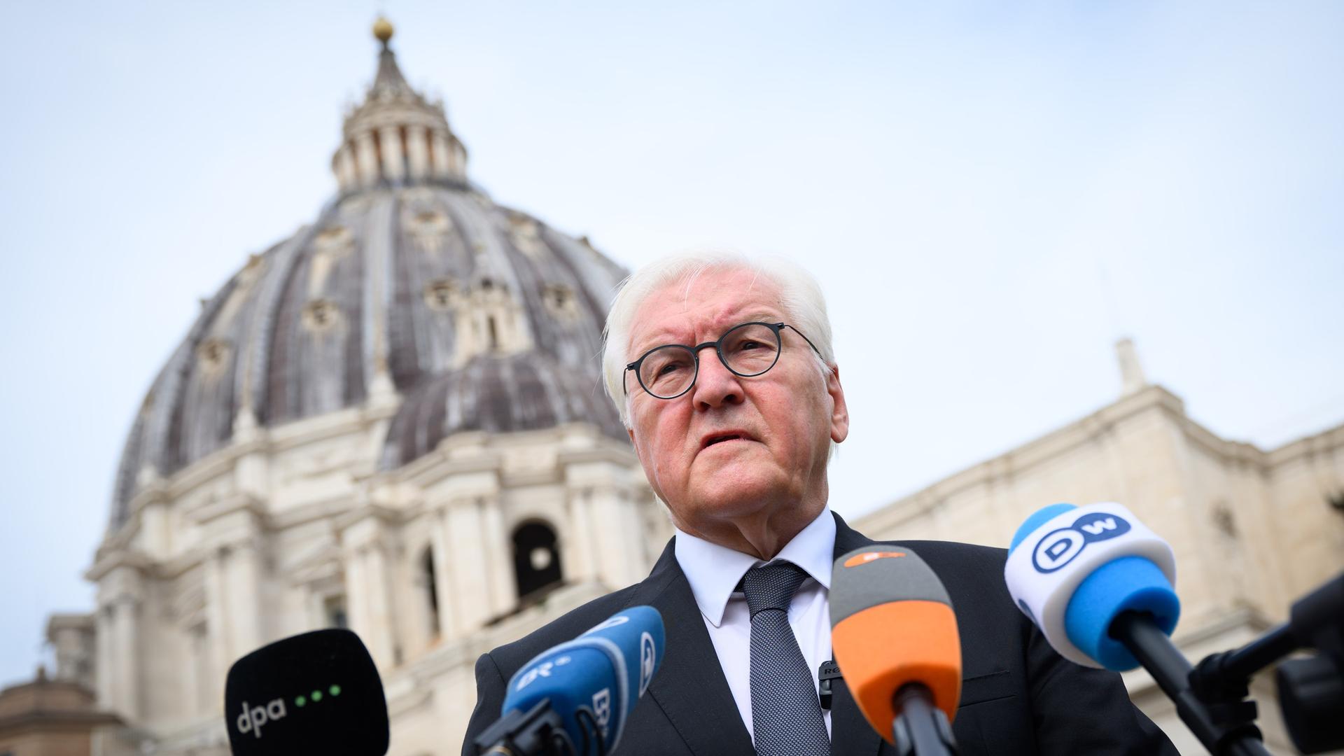 Steinmeier steht vor mehreren Mikrofonen von Sendern. Man sieht seinen Oberkörper aus einer tiefen Perspektive. Im Hintergrund unscharf die Kuppel des Petersdoms.