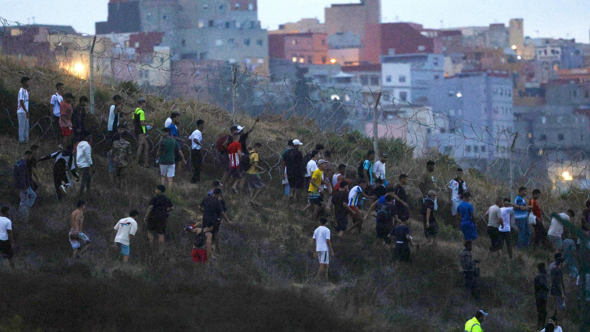 Menschen sammeln sich auf einem Hügel neben der Grenze zu Ceuta. Im Hintergrund sind die Gebäude der Enklave zu sehen. Menschen sammeln sich auf einem Hügel neben der Grenze zu Ceuta. Im Hintergrund sind die Gebäude der Enklave zu sehen.