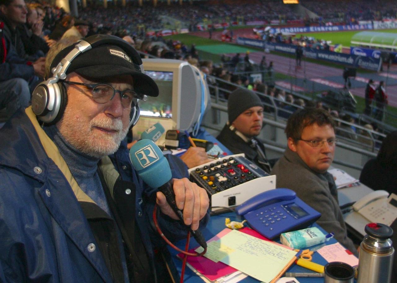 Günther Koch sitzt mit Kopfhörern und Mikrofon auf der Pressetribüne im Stadion
