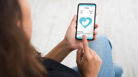 Eine junge Frau in den USA hält ihr Mobiltelefon mit einer Zyklus-App in der Hand, die Illustration von einem Fötus ist auf dem Display zu sehen.