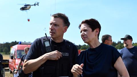 Silke Gorißen (rechts im Bild), Ministerin für Landwirtschaft und Verbraucherschutz des Landes Nordrhein-Westfalen, steht mit einem Polizeibeamten vor der Einsatzzentrale beim Waldbrand im Hürtgenwald. Im Hintergrund fliegt ein Löschhubschrauber vorbei.
