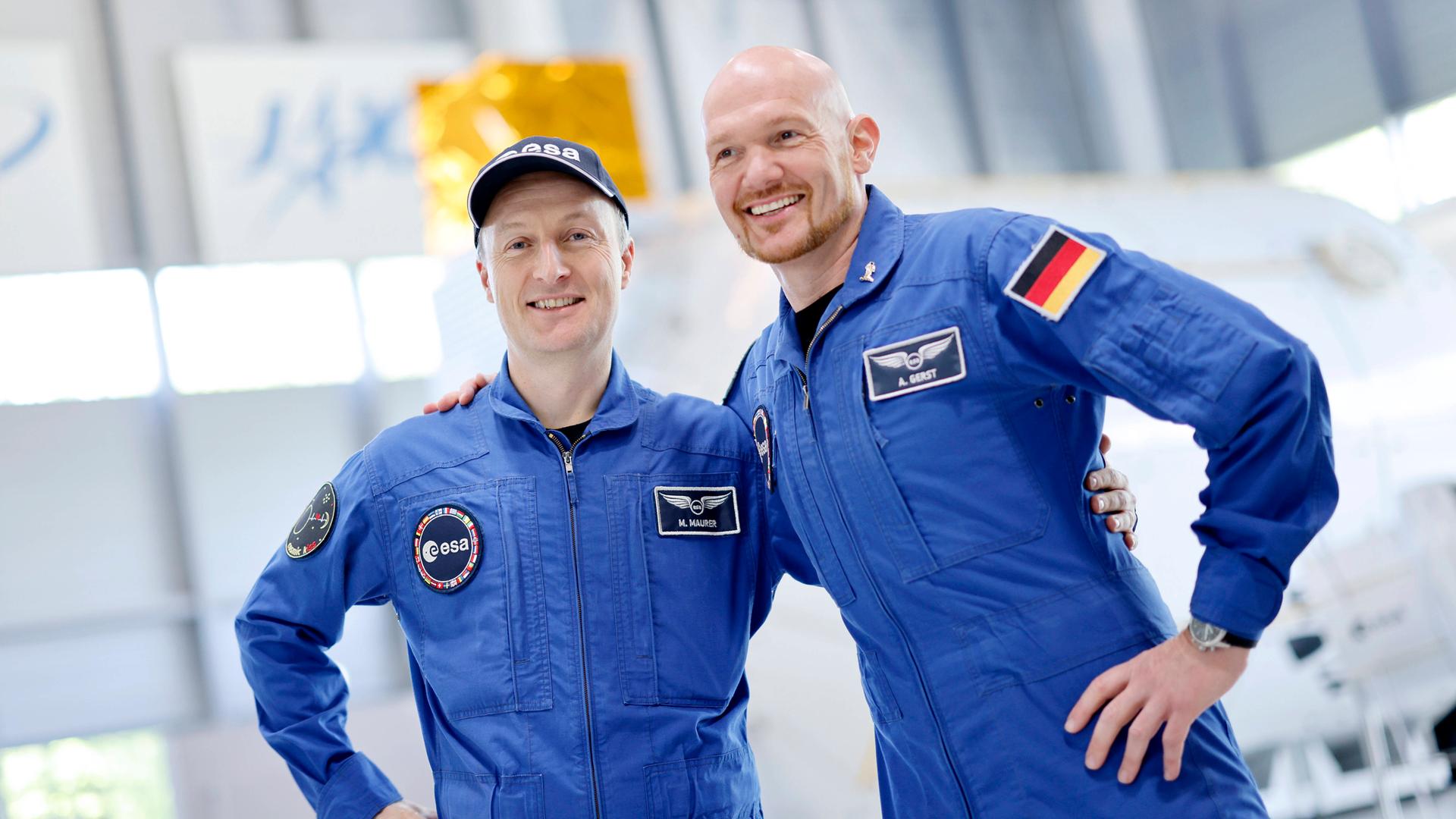Die deutschen Astronauten Matthias Maurer (li.) und Alexander Gerst (re.) stehen Arm in Arm in blauen Anzügen. Die deutschen Astronauten Matthias Maurer (li.) und Alexander Gerst (re.) stehen Arm in Arm in blauen Anzügen.