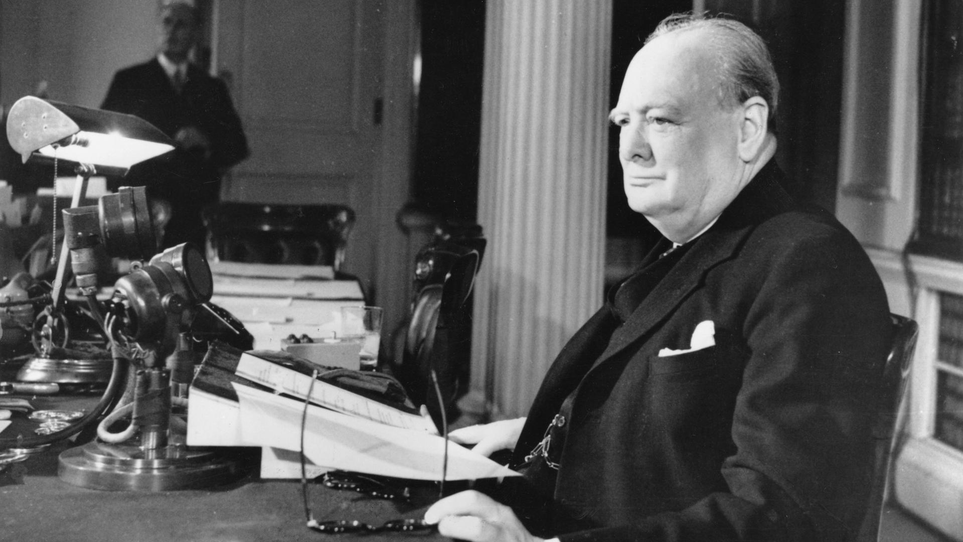Winston Churchill sitzt mit einer Zeitung an einem Schreibtisch