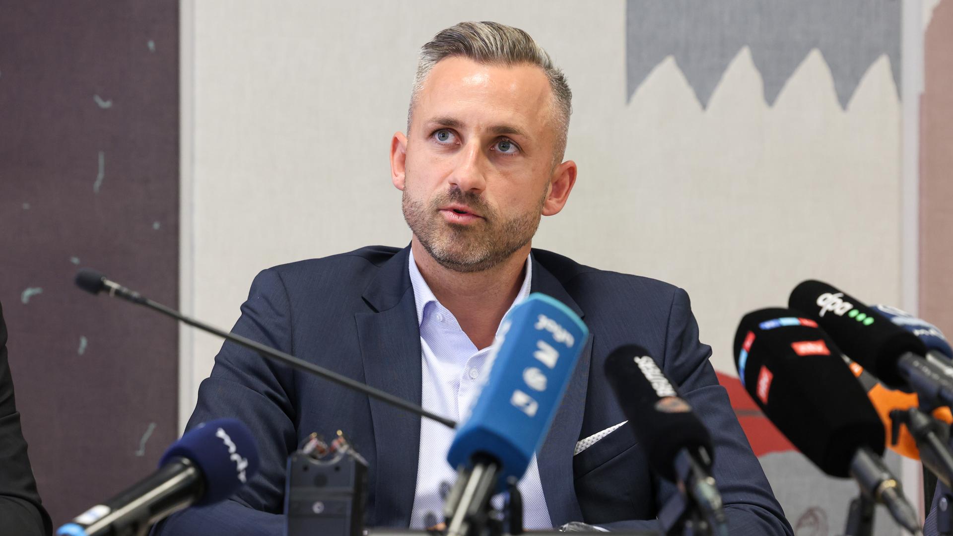 Siegmund sitzt an einem Tisch vor mehreren Mikrofonen und sagt etwas. Er trägt ein dunkles Jackett und ein hellblaues Hemd. Man sieht ihn von vorne. Siegmund sitzt an einem Tisch vor mehreren Mikrofonen und sagt etwas. Er trägt ein dunkles Jackett und ein hellblaues Hemd. Man sieht ihn von vorne.