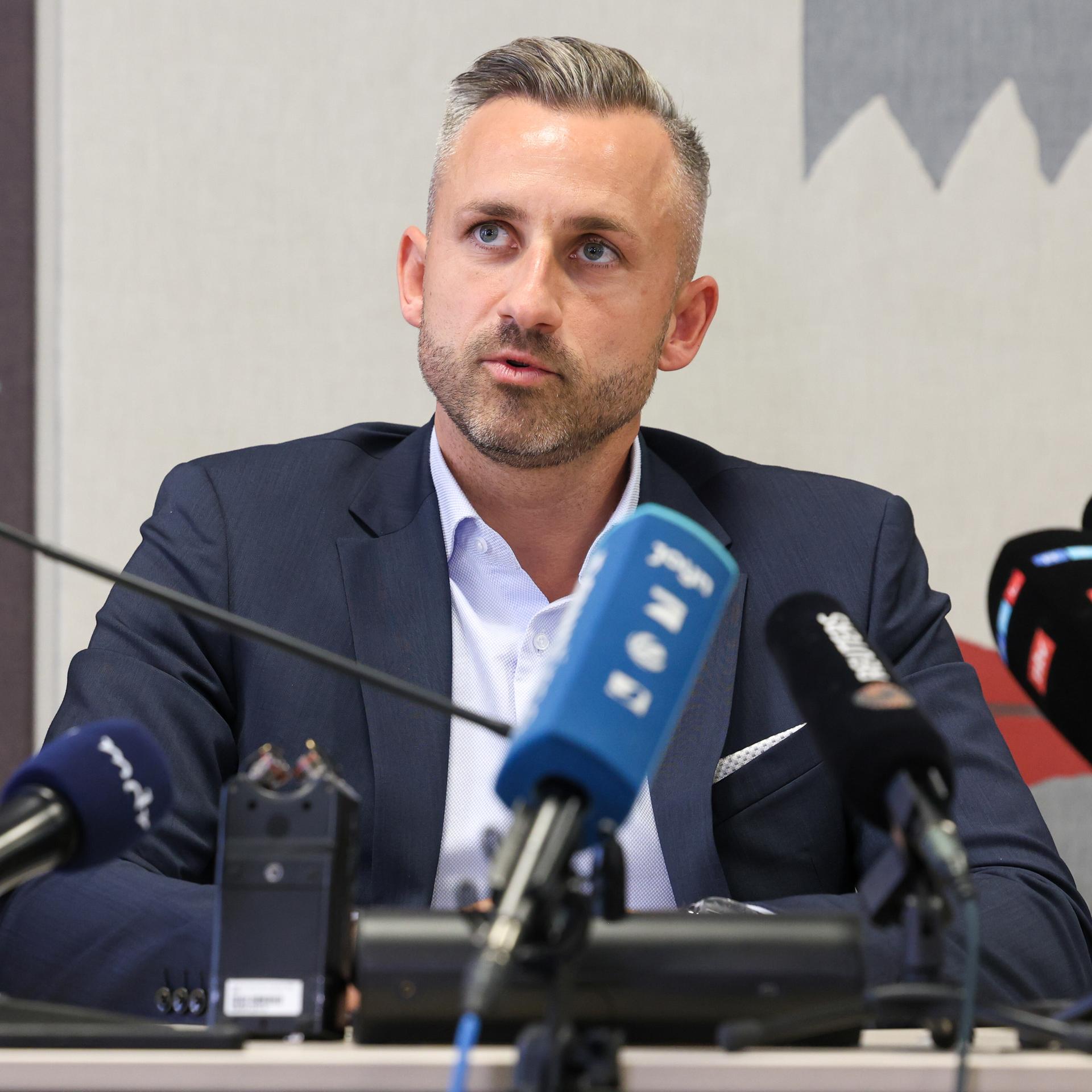 Siegmund sitzt an einem Tisch vor mehreren Mikrofonen und sagt etwas. Er trÀgt ein dunkles Jackett und ein hellblaues Hemd. Man sieht ihn von vorne. Siegmund sitzt an einem Tisch vor mehreren Mikrofonen und sagt etwas. Er trÀgt ein dunkles Jackett und ein hellblaues Hemd. Man sieht ihn von vorne.