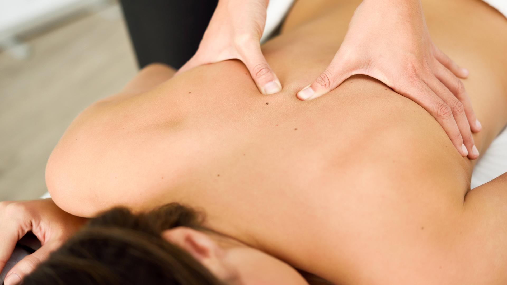 Zwei Hände massieren den Rücken einer Frau, die auf dem Bauch einer Massageliege liegt.