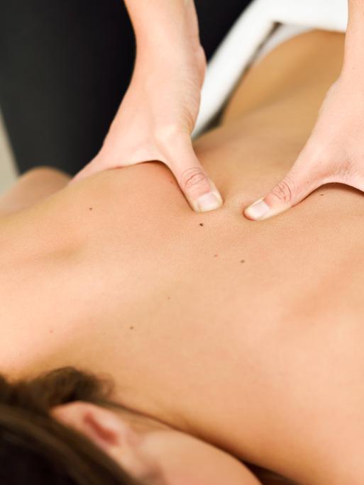 Zwei Hände massieren den Rücken einer Frau, die auf dem Bauch einer Massageliege liegt.