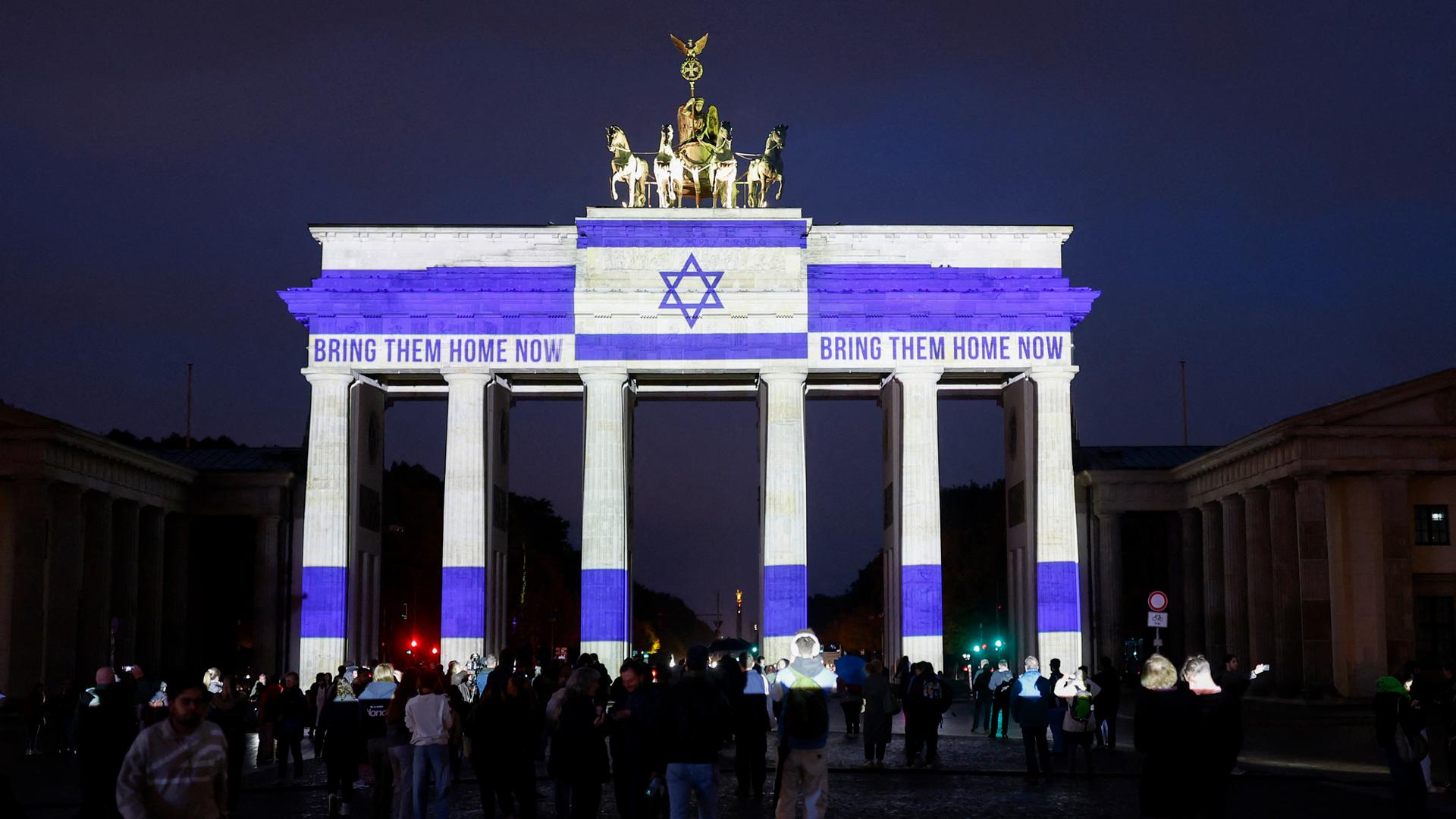Nachtaufnahme des Brandenburger Tors, das mit der israelischen Flagge und den Worten "Bring them Home" angestrahlt wird. Davor sind zahlreiche Menschen versammelt.