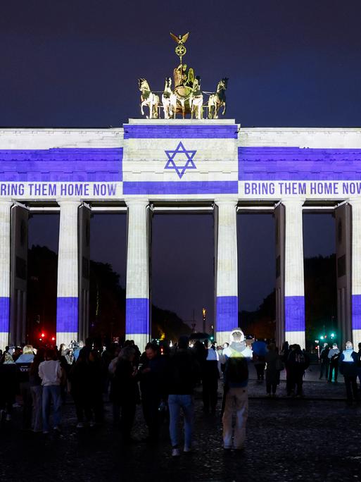 Nachtaufnahme des Brandenburger Tors, das mit der israelischen Flagge und den Worten "Bring them Home" angestrahlt wird. Davor sind zahlreiche Menschen versammelt. Nachtaufnahme des Brandenburger Tors, das mit der israelischen Flagge und den Worten "Bring them Home" angestrahlt wird. Davor sind zahlreiche Menschen versammelt.