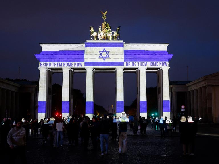 Nachtaufnahme des Brandenburger Tors, das mit der israelischen Flagge und den Worten "Bring them Home" angestrahlt wird. Davor sind zahlreiche Menschen versammelt. Nachtaufnahme des Brandenburger Tors, das mit der israelischen Flagge und den Worten "Bring them Home" angestrahlt wird. Davor sind zahlreiche Menschen versammelt.