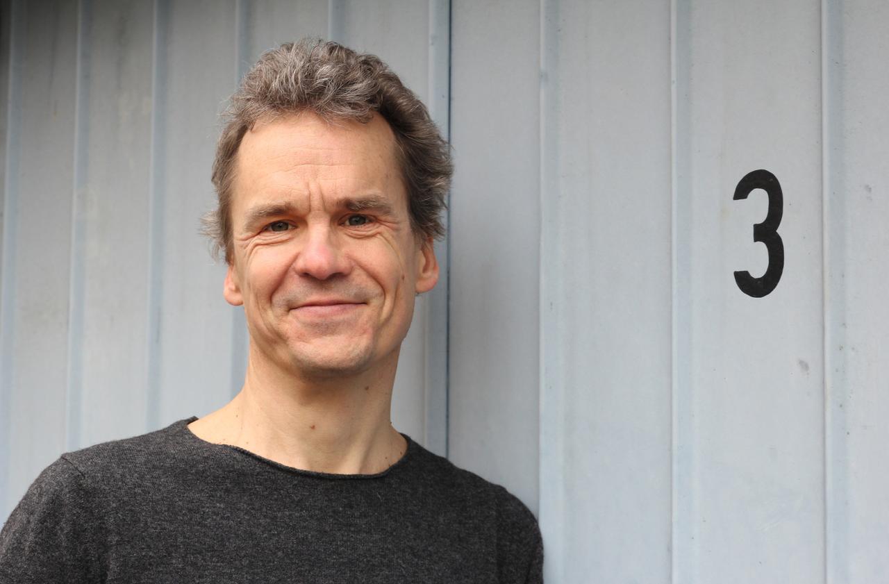 Porträtfoto des Journalisten und Autors Alex Rühle. Er trägt einen dunklen Pullover und steht vor einer hellgrauen Holzwand.