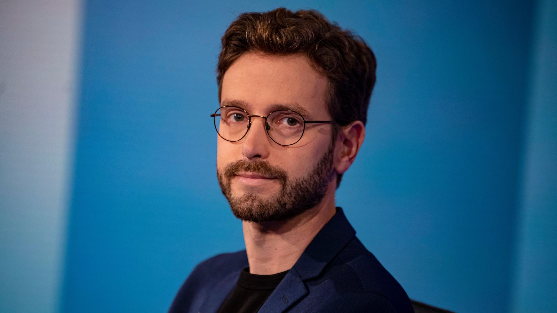 Tobias Bank (Die Linke), Bundesgeschäftsführer, schaut vor einem blauen Hintergrund in die Kamera.