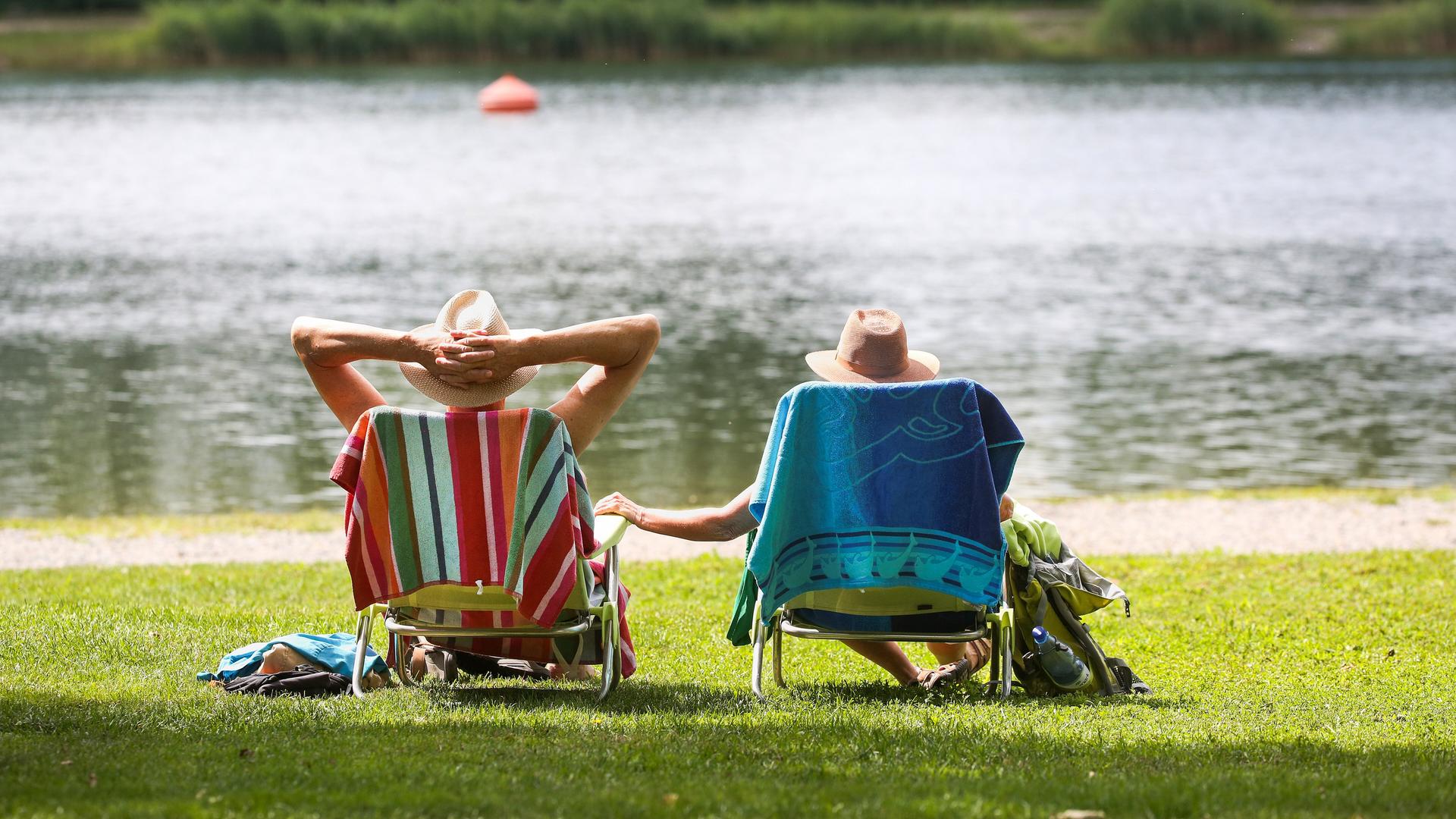 Ein Mann und eine Frau mit Strohhüten sitzen an einem Badesee in ihren Stühlen in der Sonne.