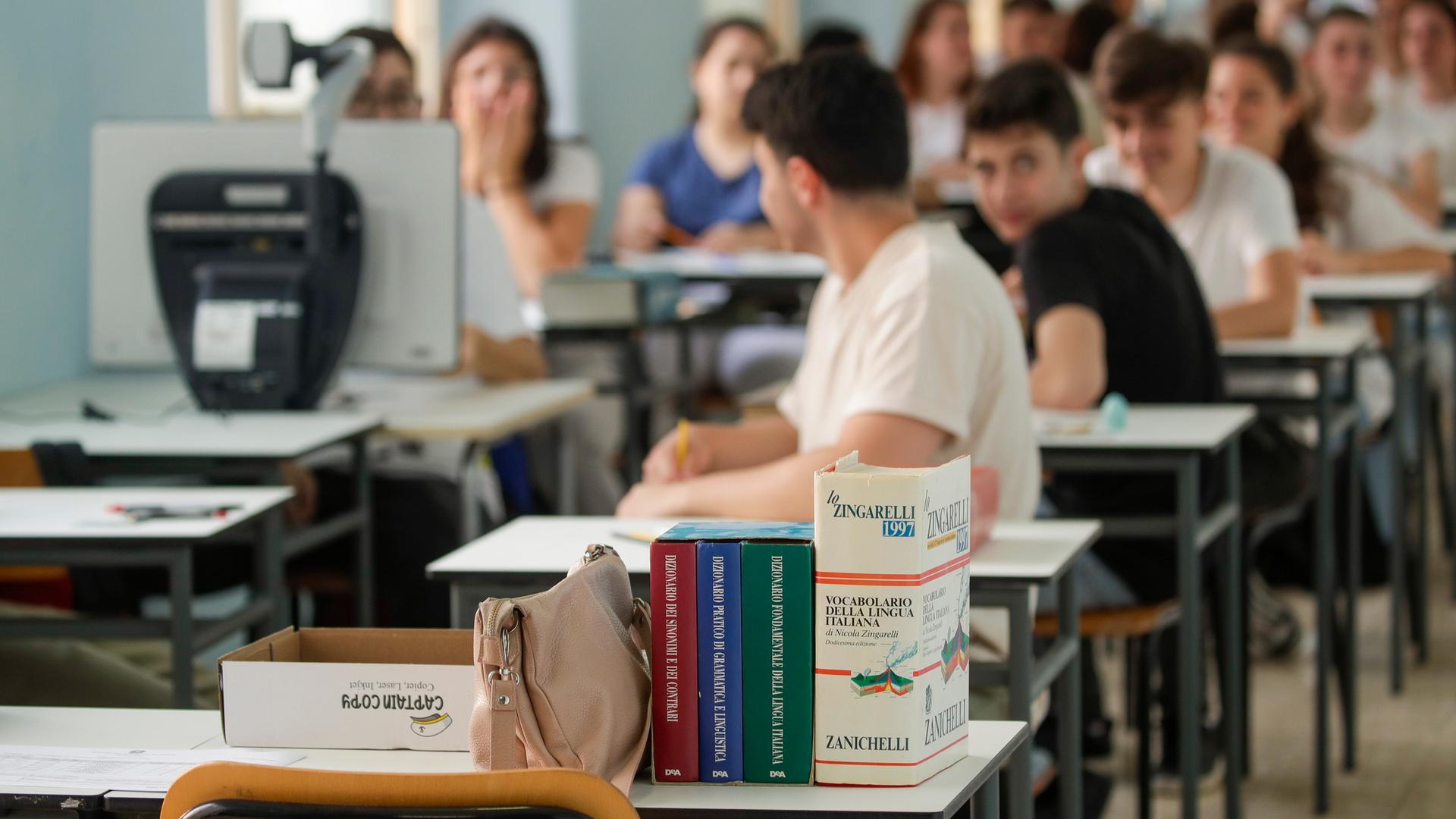 Jugendliche sitzen in einem Klassenzimmer in zwei Reihen hintereinander. Auf dem ersten Tisch steht ein italienisches Schulbuch.