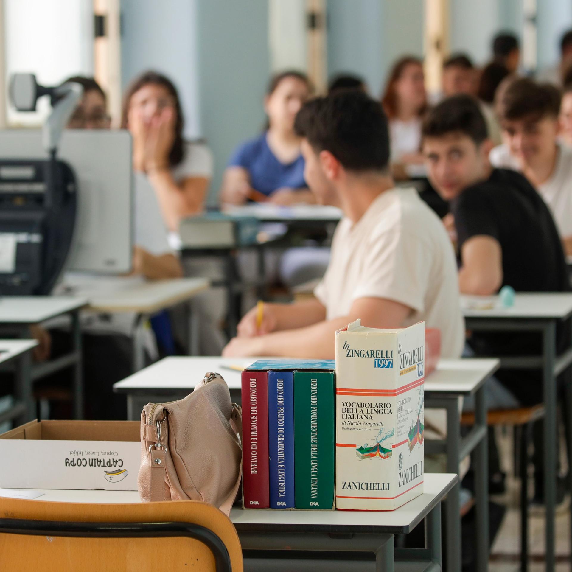 Jugendliche sitzen in einem Klassenzimmer in zwei Reihen hintereinander. Auf dem ersten Tisch steht ein italienisches Schulbuch.