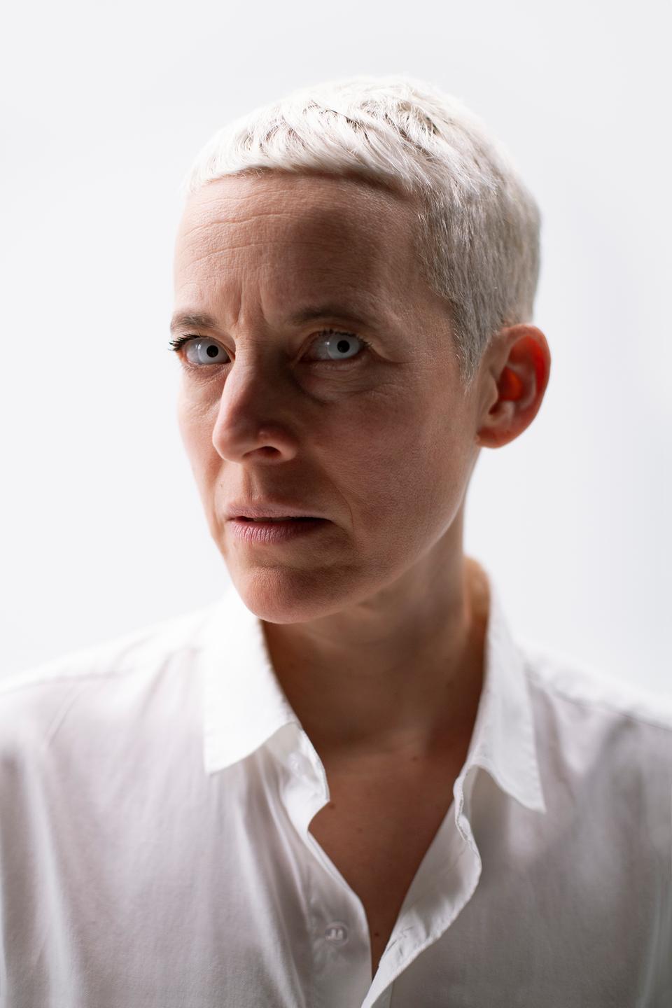 Die Künstlerin Candice Breitz im weißen Hemd mit blondem Kurzhaarschnitt schaut mit wasserhellen Augen frontal in die Kamera.