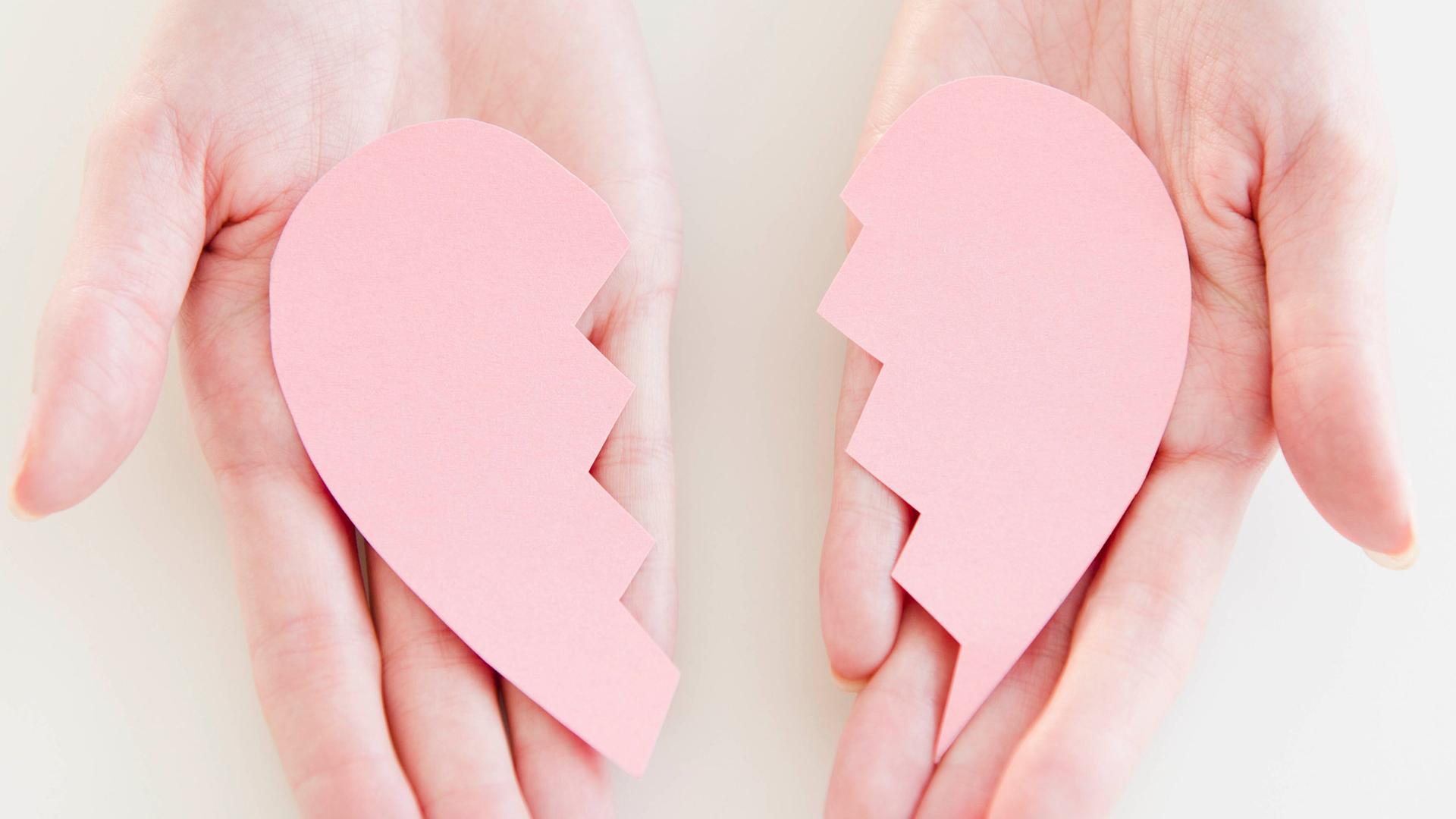 Ein rosafarbenes zerbrochenes Herz aus Kunststoff liegt auf einem Paar Handflächen