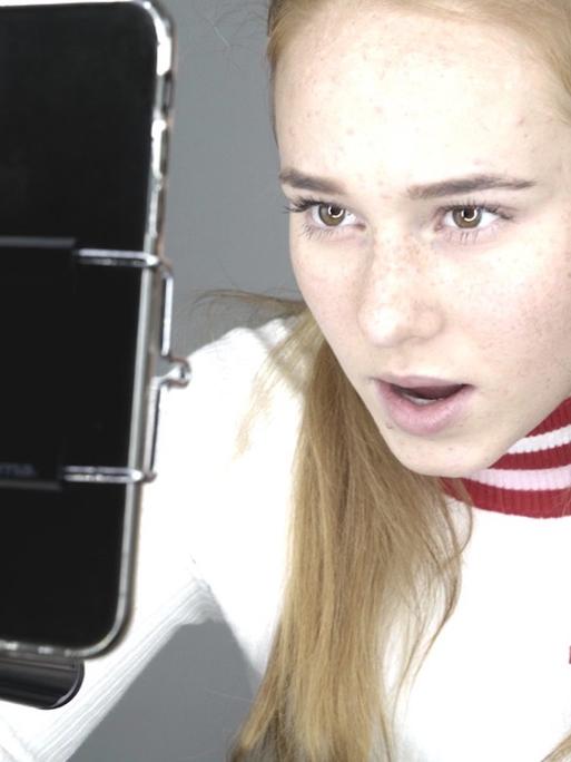 Im Still aus "Girl Gang" spricht ein Mädchen mit langem blonden Haar in die Selfiekamera ihres Smartphones.