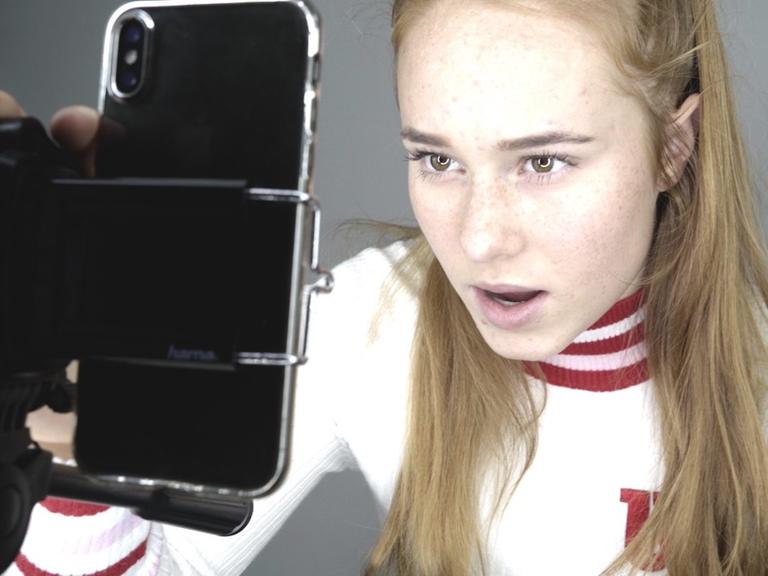 Im Still aus "Girl Gang" spricht ein Mädchen mit langem blonden Haar in die Selfiekamera ihres Smartphones.