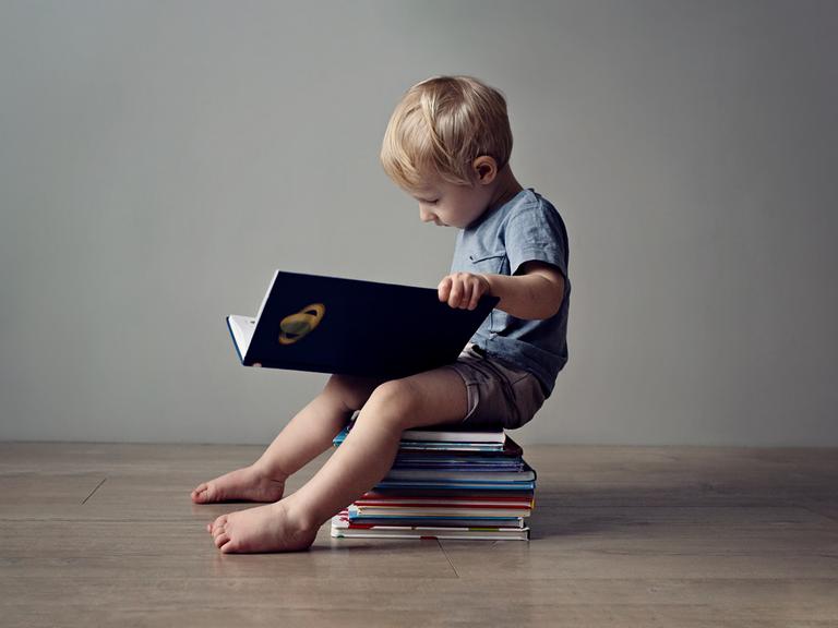 Ein kleiner Junge mit BUch in der Hansd sitzt auf einem Bücherstapel und liest