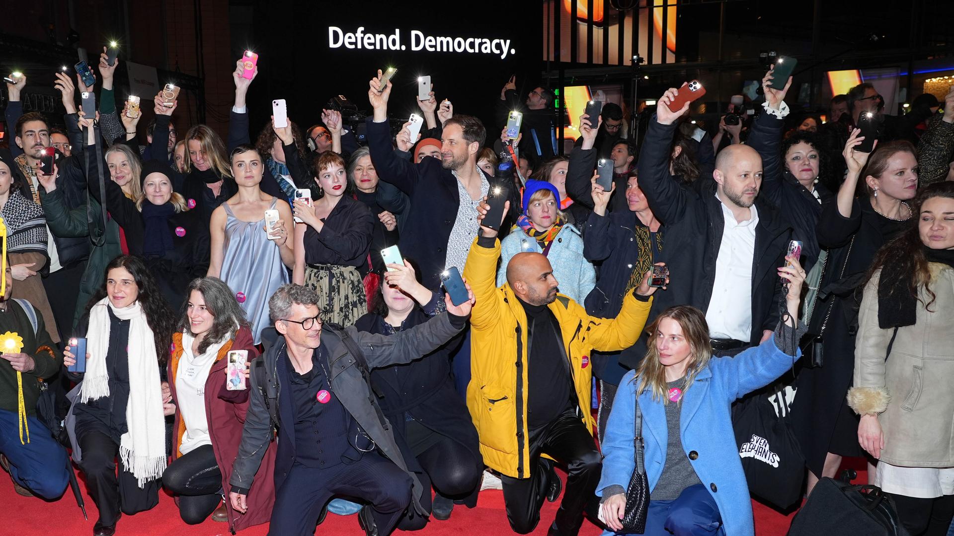 Eine Gruppe Schauspieler hält ihre Handys in die Luft. Im Hintergrund ist der Schriftzug "Defend Democracy" zu lesen.