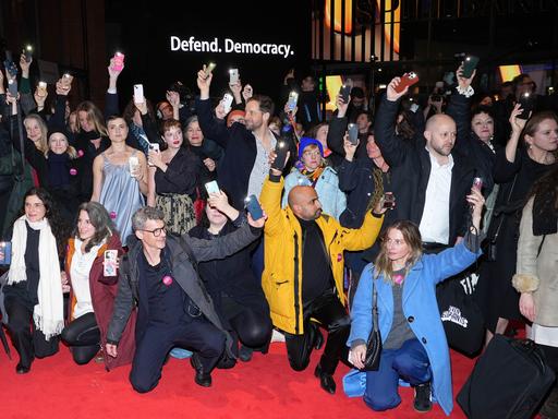 Eine Gruppe Schauspieler hält ihre Handys in die Luft. Im Hintergrund ist der Schriftzug "Defend Democracy" zu lesen.