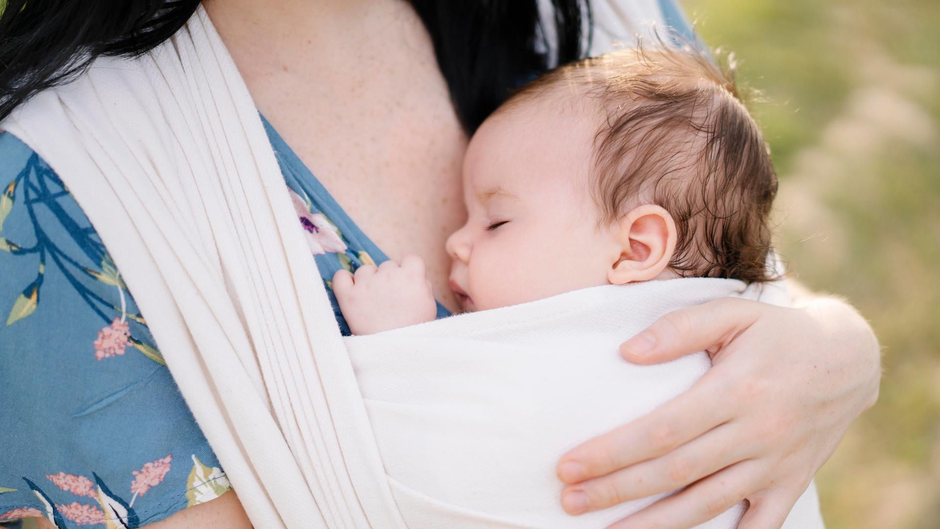Eine Mutter trägt ein schlafendes Baby, das in einem Tuch gewickelt ist. 