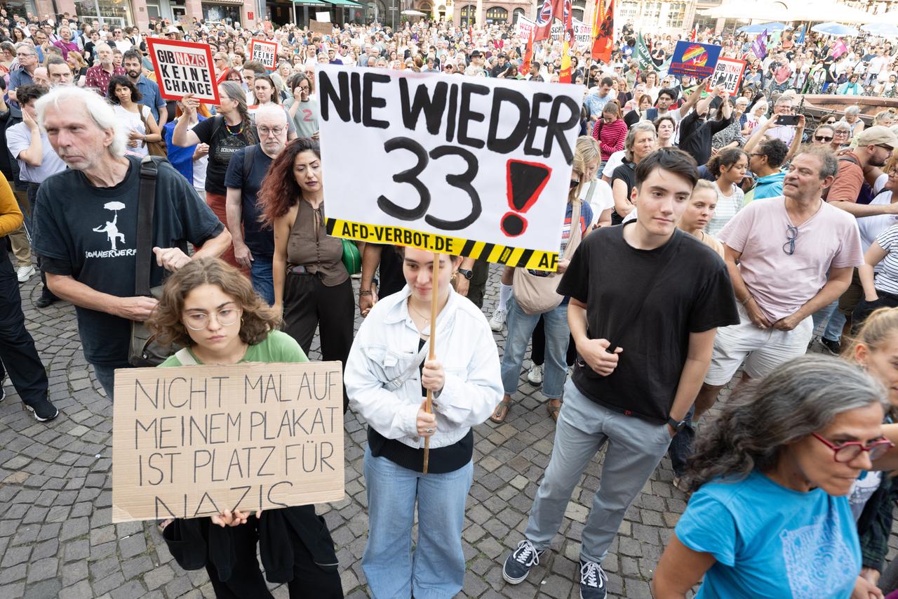 Viele Menschen stehen auf einem Platz, einige halten Plakate und Banner hoch. Auf einem Plakat weit vorne steht "Nie wieder 33".