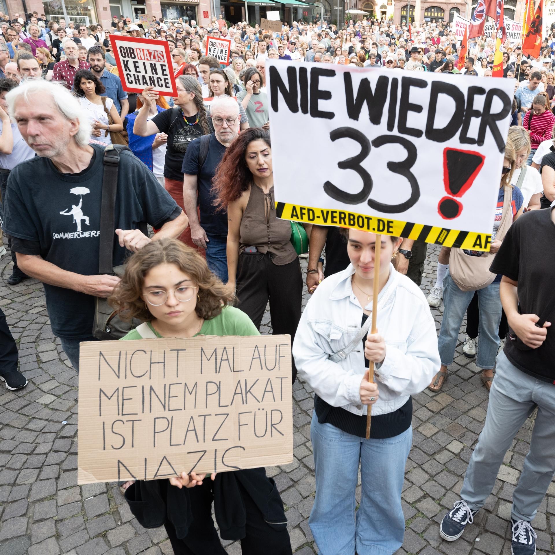 Viele Menschen stehen auf einem Platz, einige halten Plakate und Banner hoch. Auf einem Plakat weit vorne steht "Nie wieder 33".