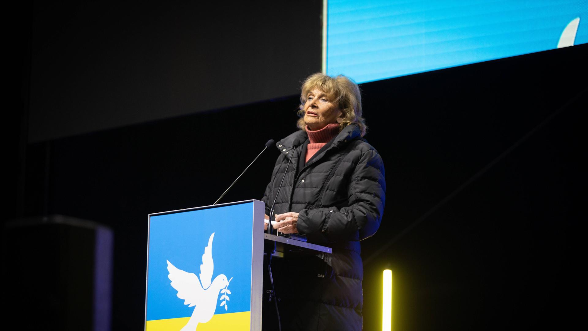 Die ehemalige Präsidentin des Zentralrats der Juden in Deutschland, Charlotte Knobloch, steht hinter einem Rednerpult in Farben der ukrainischen Flagge und spricht in ein Mikrofon.