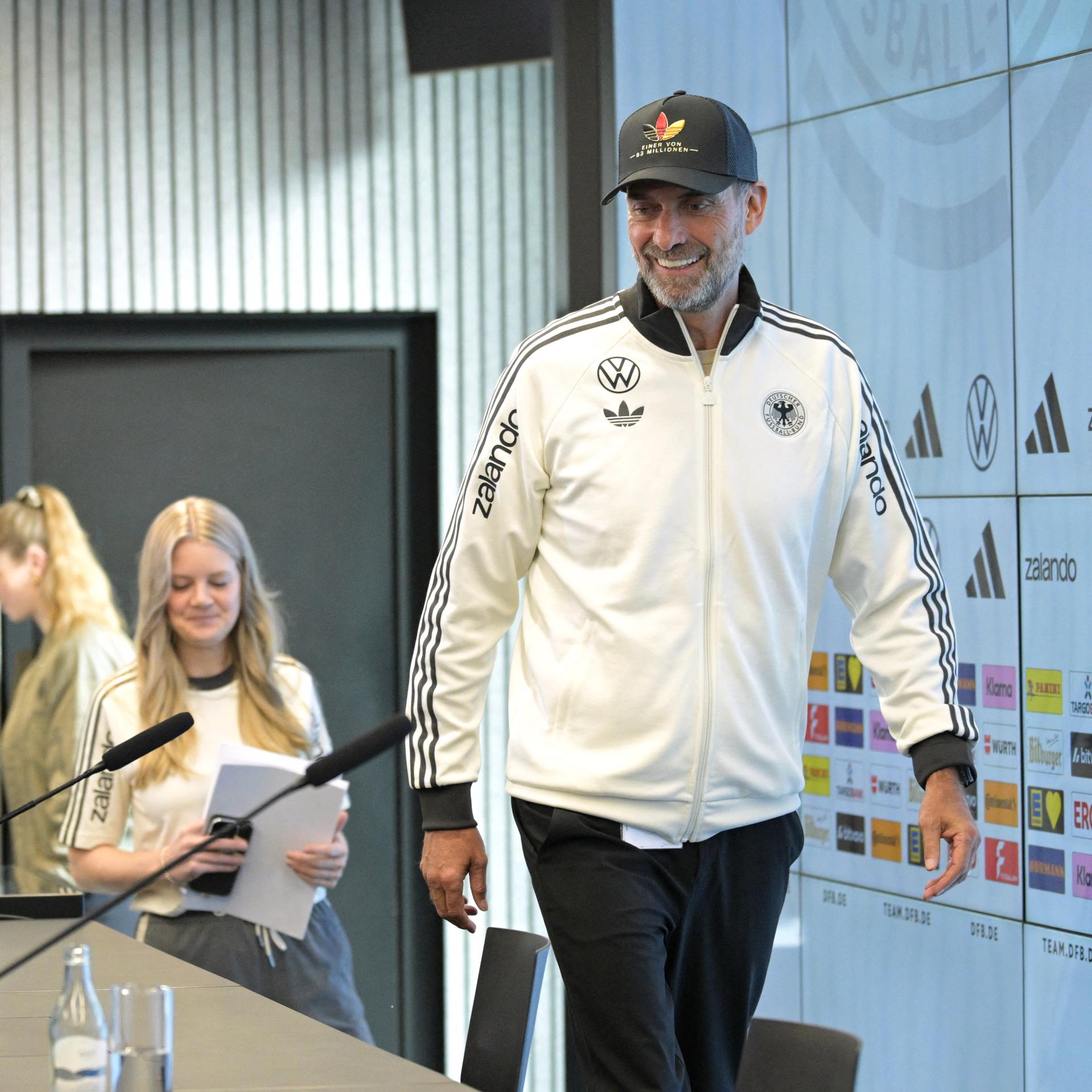 Ein Mann mit Kappe betritt eine Bühne zur Pressekonferenz.