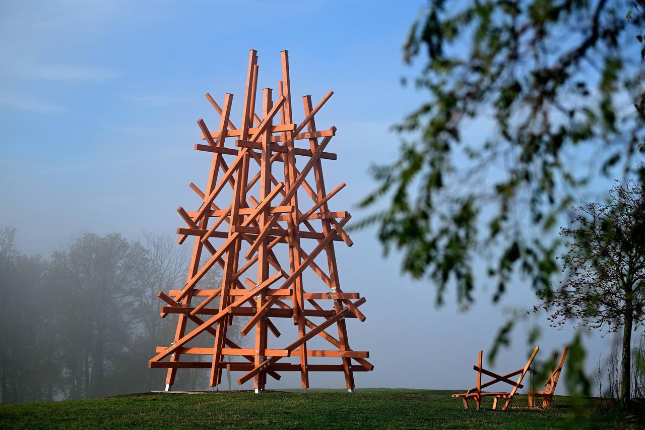 Eine Skulptur aus brunen Stäben ragt über einer grünen Wiese in den blauen Himmel