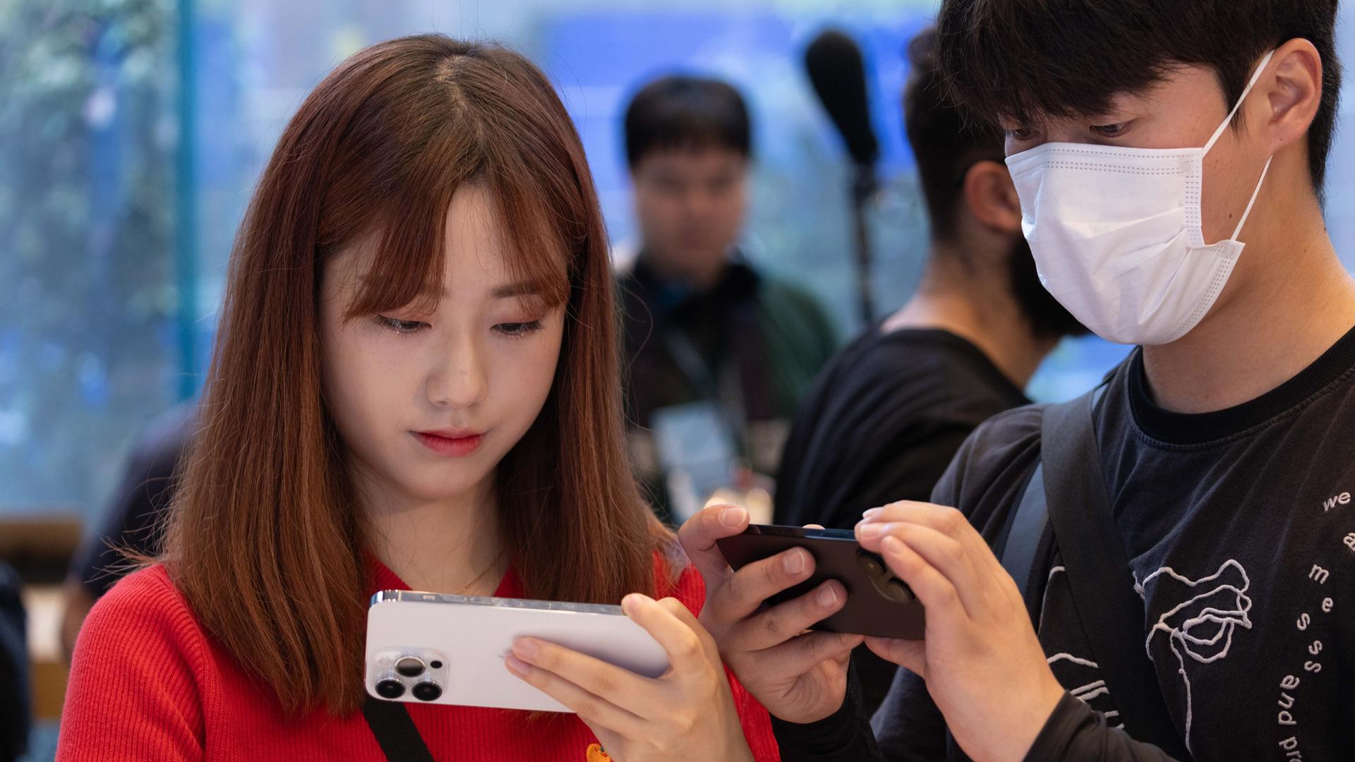 Ein junges Mädchen und ein junger Mann in Japan schauen auf die Displays ihrer Mobiltelefone, der Jugendliche trägt einen Mund-Nasen-Schutz.