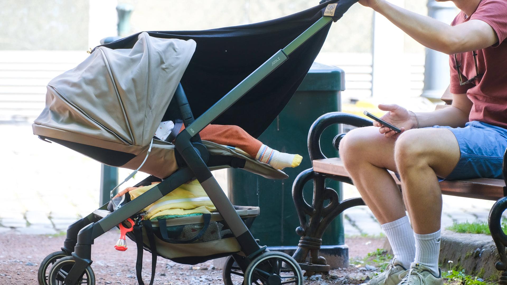 Ein Mann sitzt auf einer Bank und hält einen Kinderwagen fest, in dem ein Kleinkind liegt.