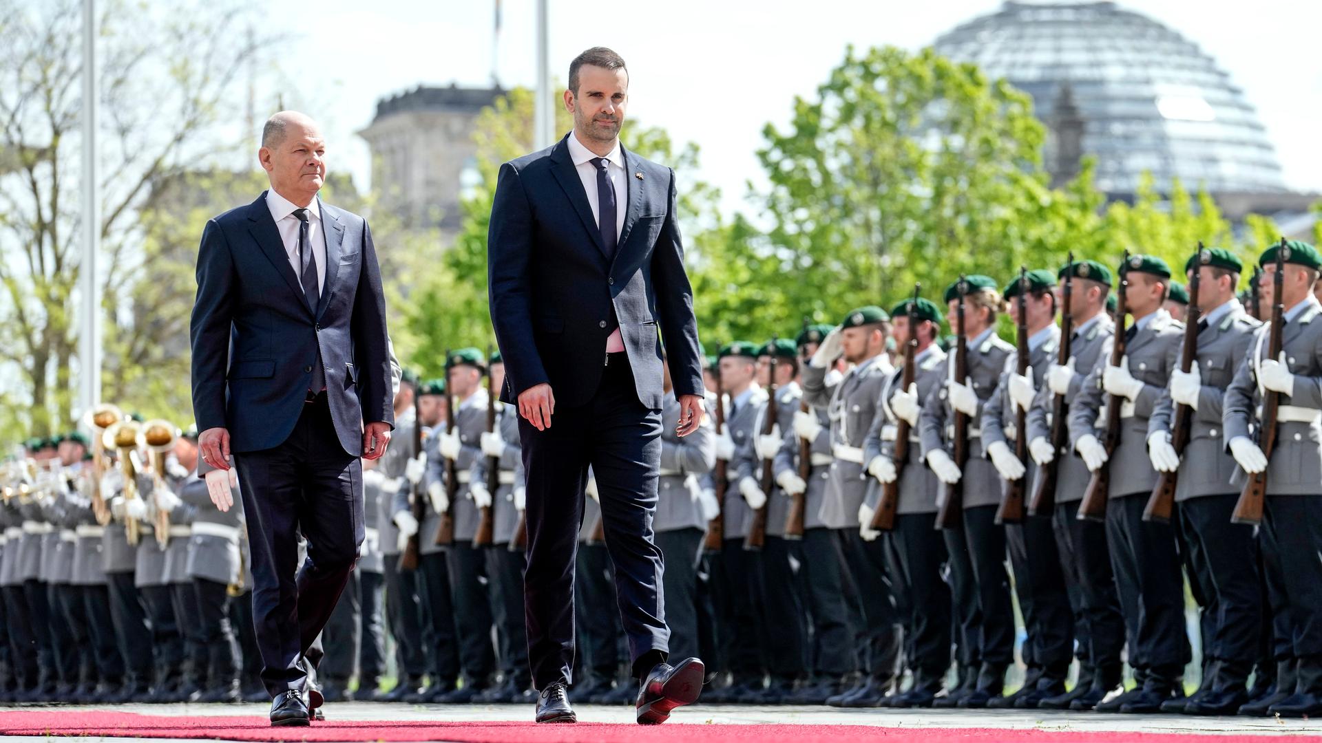 Bundeskanzler Olaf Scholz (links) und Montenegros Ministerpräsident Milojko Spajic schreiten vor Soldatinnen und Soldaten einen roten Teppich entlang. Dahinter die Kuppel des Reichstags. 