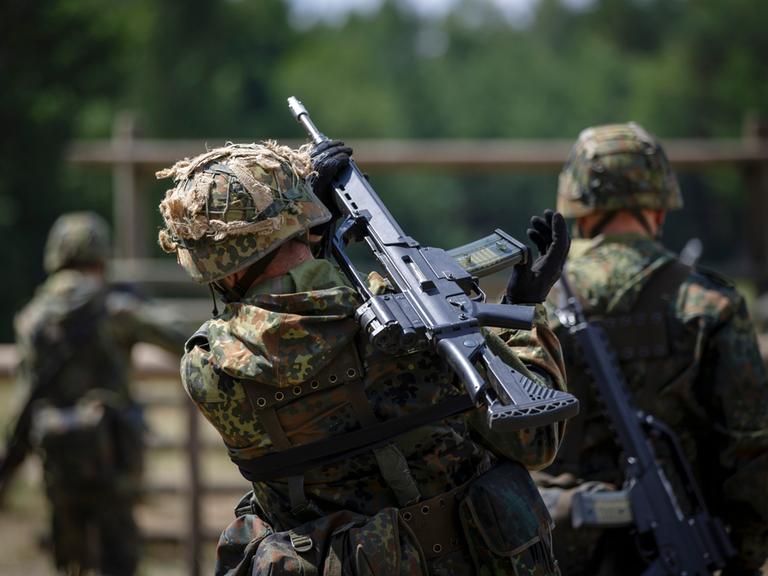 Drei Rekruten marschieren auf einem Gelände, sie haben Gewehre geschultert. Alle drei tragen Tarnanzüge und sind von hinten zu sehen.