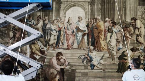 Detail der Anbringung eines Wanteppichs. Links im Bild ist eine Hebebühne zu sehen, zwei Arbeiter stützen den Wanteppich mit Stangen.
Das Motiv zeigt einen monumentalen Innenraum, in dem eine Versammlung maßgeblicher Wissenschaftler und Philosophen von der Antike bis zur Renaissance stattfindet. 
