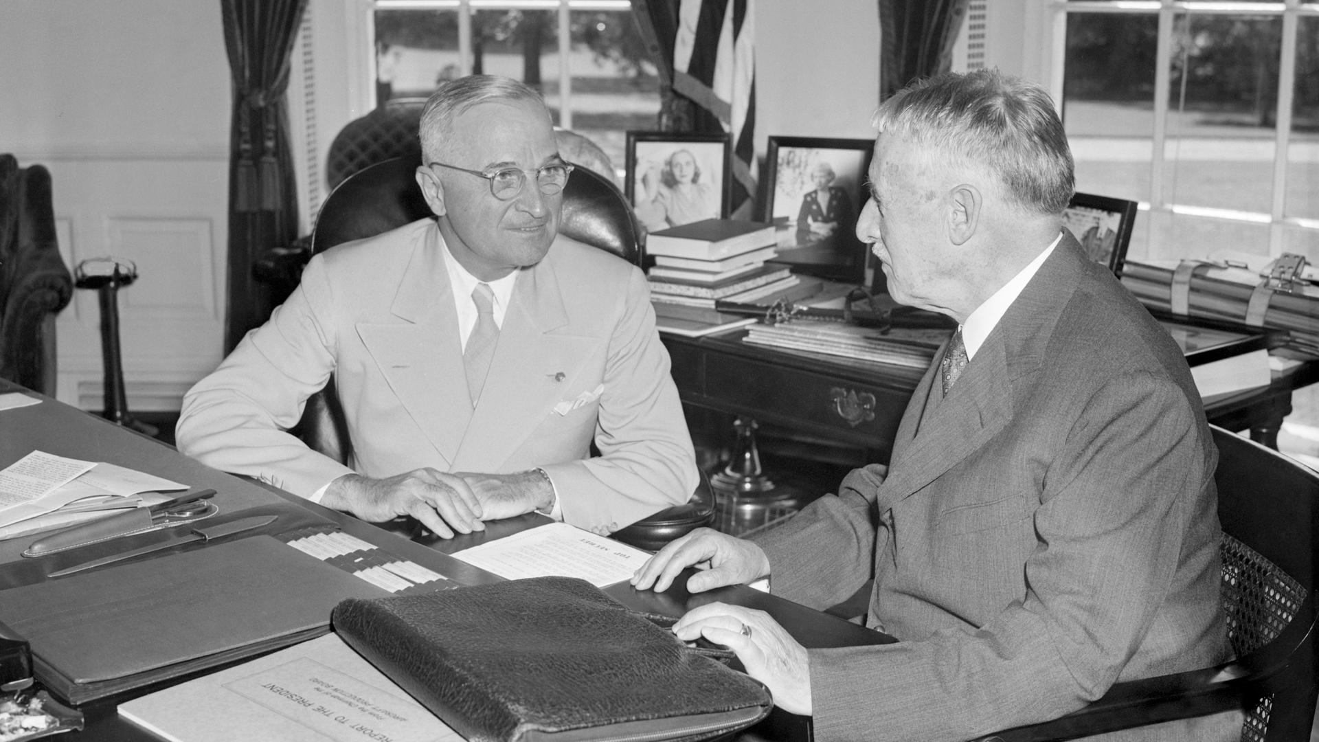 Harry Truman und Henry Stimson sitzen in Trumans Büro am Schreibtisch. Sie sind einander zugewandt und unterhalten sich.