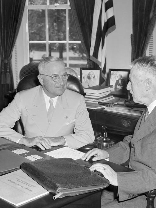Harry Truman und Henry Stimson sitzen in Trumans Büro am Schreibtisch. Sie sind einander zugewandt und unterhalten sich.