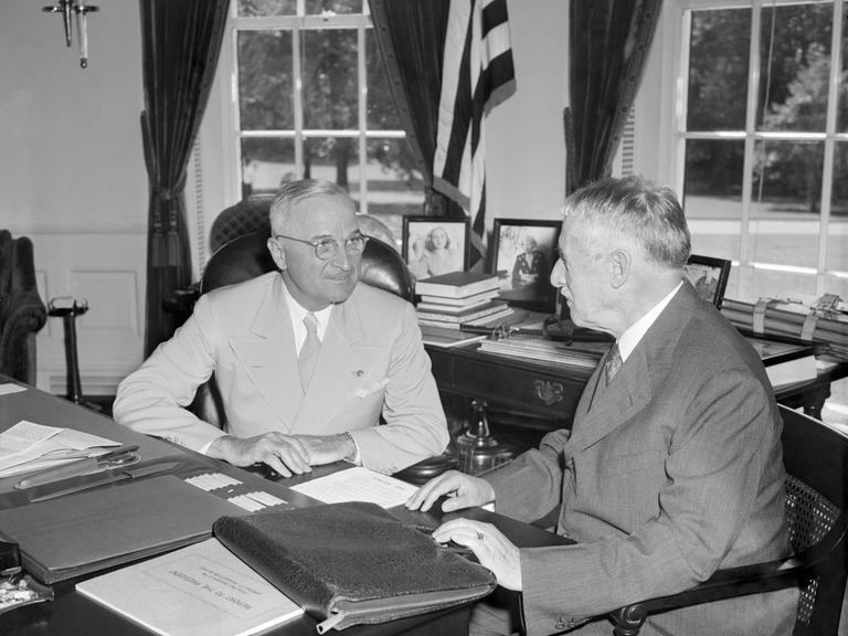 Harry Truman und Henry Stimson sitzen in Trumans Büro am Schreibtisch. Sie sind einander zugewandt und unterhalten sich.