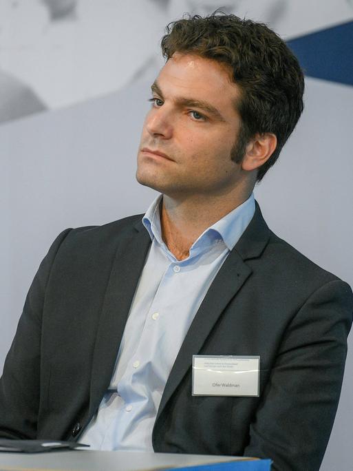 Ofer Waldman sitzt bei einer Veranstaltung auf der Bühne. Er trägt einen Anzug mit hellblauem Hemd.