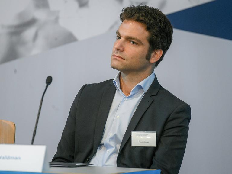 Ofer Waldman sitzt bei einer Veranstaltung auf der Bühne. Er trägt einen Anzug mit hellblauem Hemd.