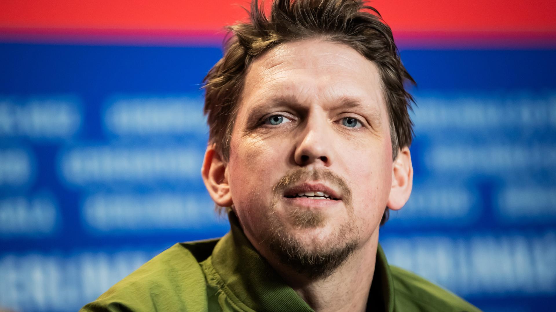 Portrait von Jan-Ole Gerster vor einem  rot-blauen Berlinale-Fotohintergrund. Er trägt ein grünes Oberteil und spricht.