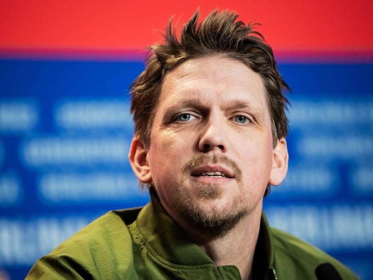 Portrait von Jan-Ole Gerster vor einem  rot-blauen Berlinale-Fotohintergrund. Er trägt ein grünes Oberteil und spricht.