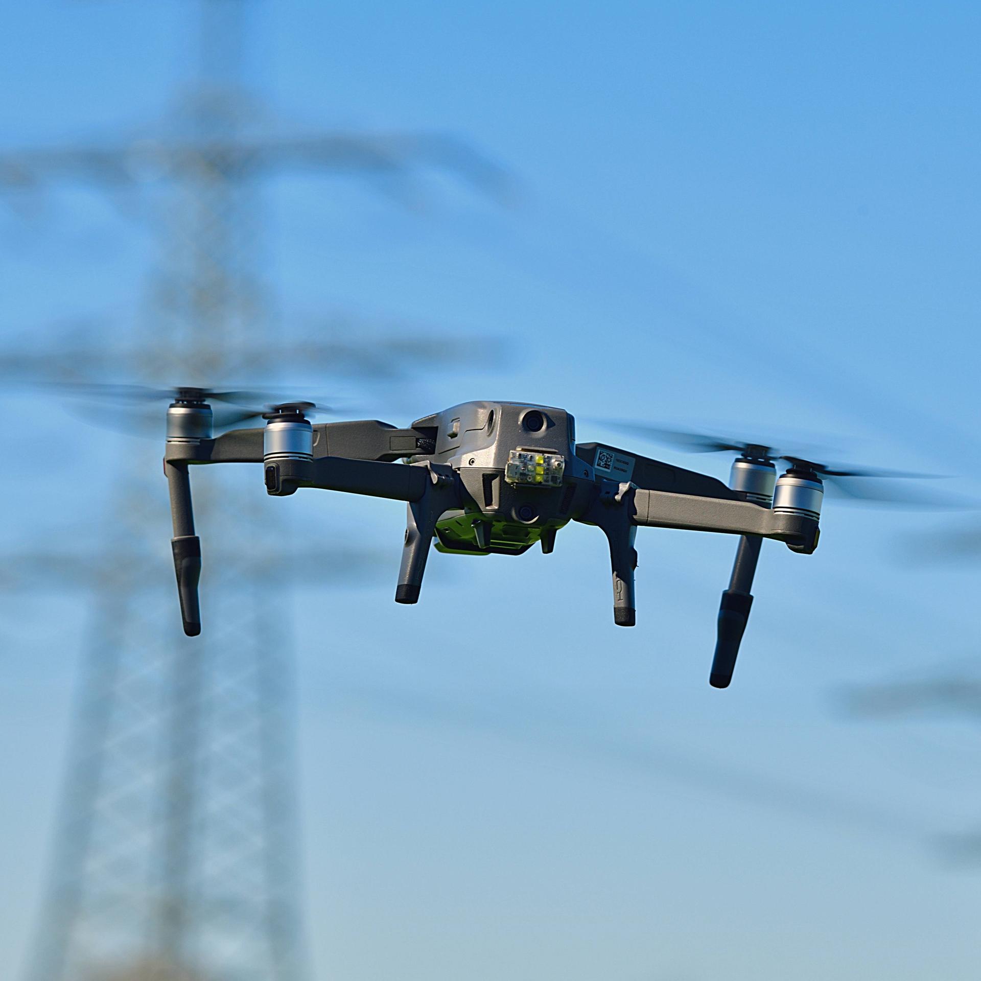 Eine graue Drohne am blauen Himmel, im Hintergrund Strommasten.