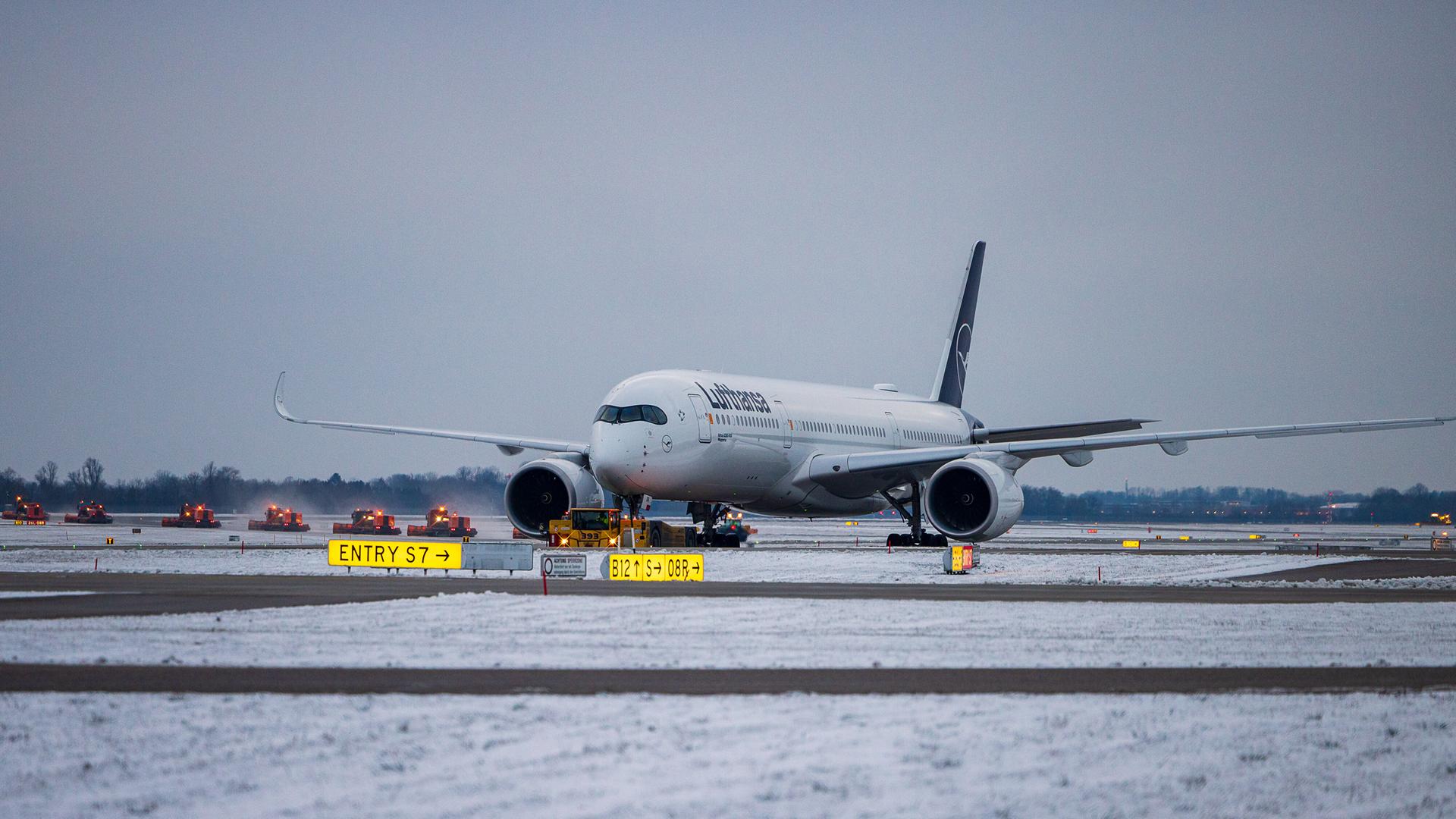 Ein Airbus A350 der Lufthansa steht am Rollweg des Flughafens Muenchen. Im Hintergrund raeumen zahlreiche Winterdienstfahrzeuge parallel Schnee von der Start- und Landebahn. 