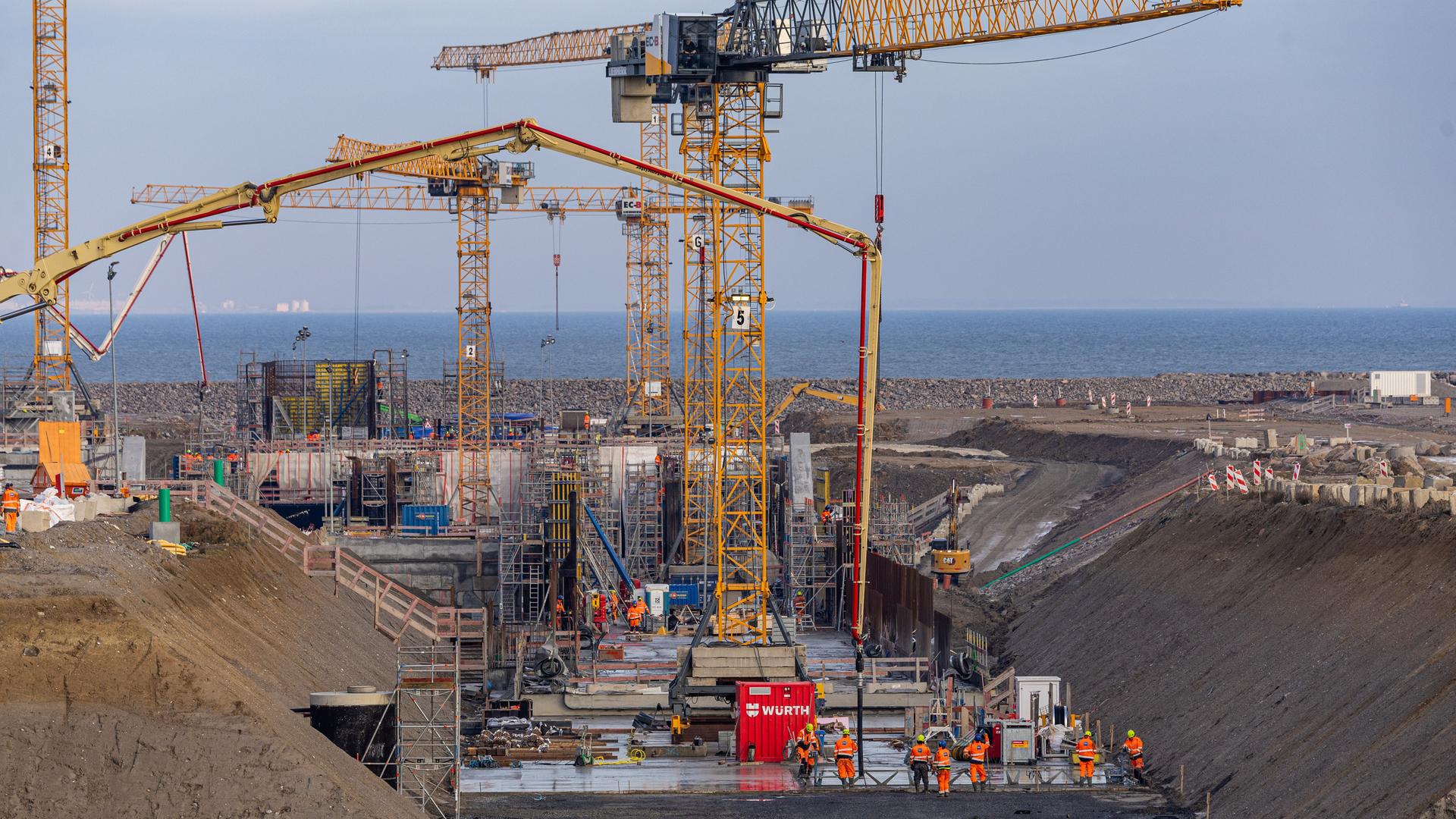 Auf einer riesigen Baustelle stehen mehrere Kräne, Menschen in Overalls arbeiten am Boden. Im Hintergrund ist das Meer zu sehen.