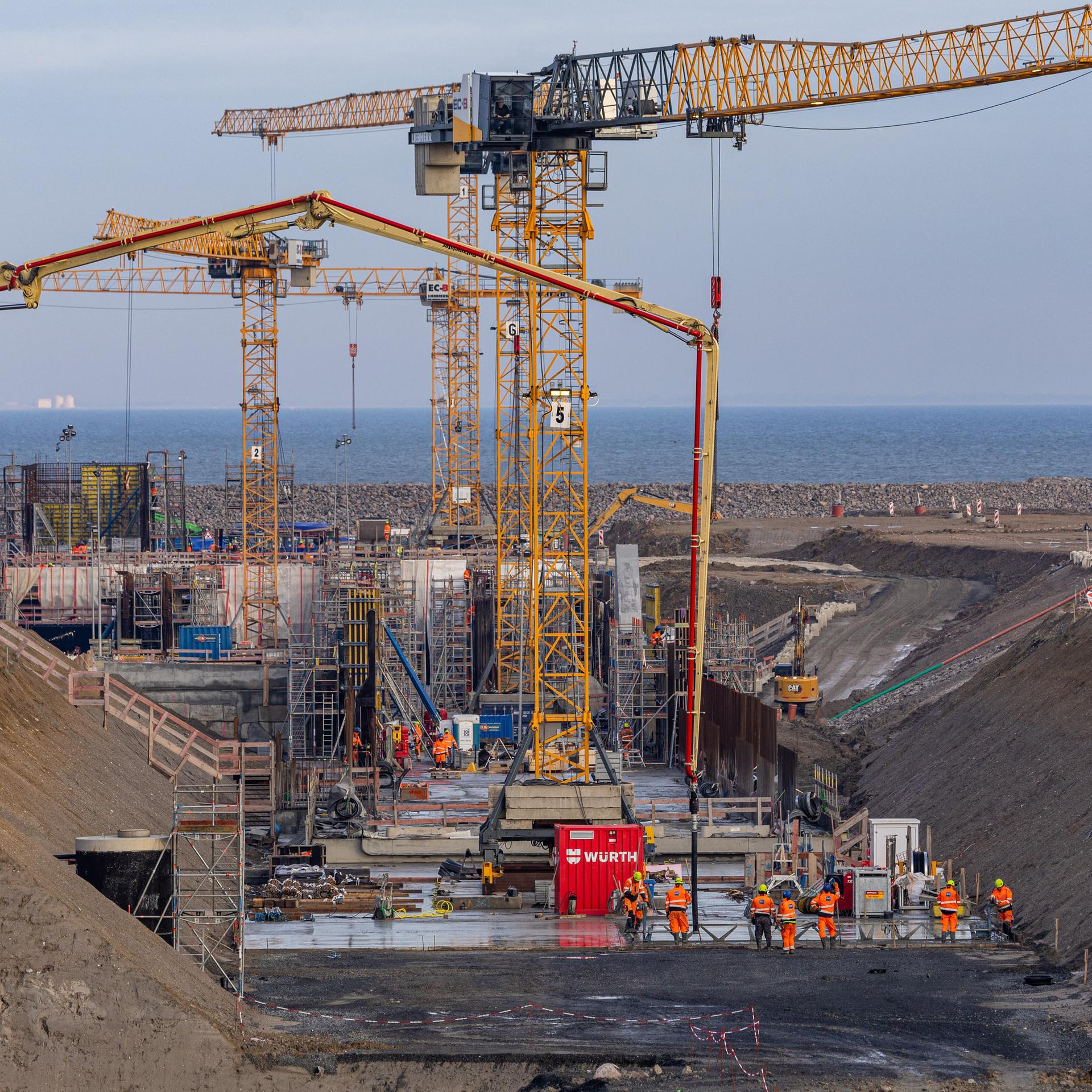 Auf einer riesigen Baustelle stehen mehrere Kräne, Menschen in Overalls arbeiten am Boden. Im Hintergrund ist das Meer zu sehen.